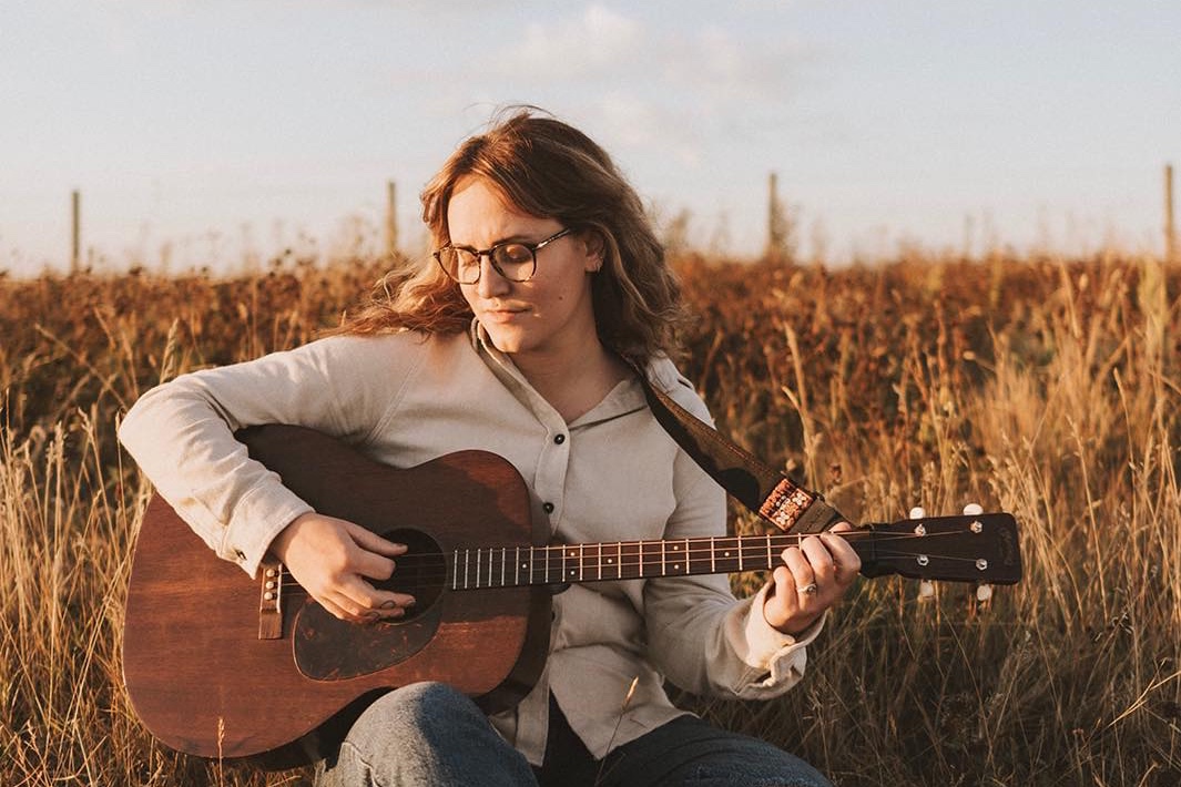 A brunette woman sits amidst tall yellow grass, playing an acoustic guitar. She wears blue jeans, a cream sweater, and glasses.