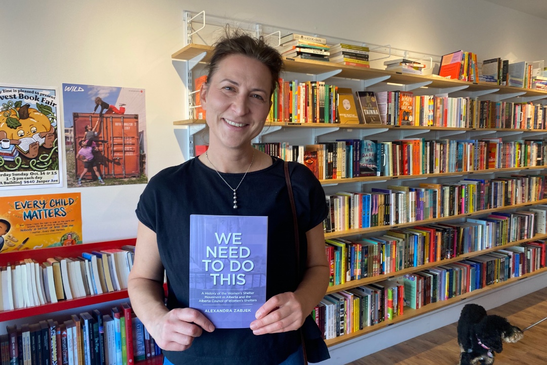 A smiling woman stands in a bookstore, holding a book titled "We Need to Do This."