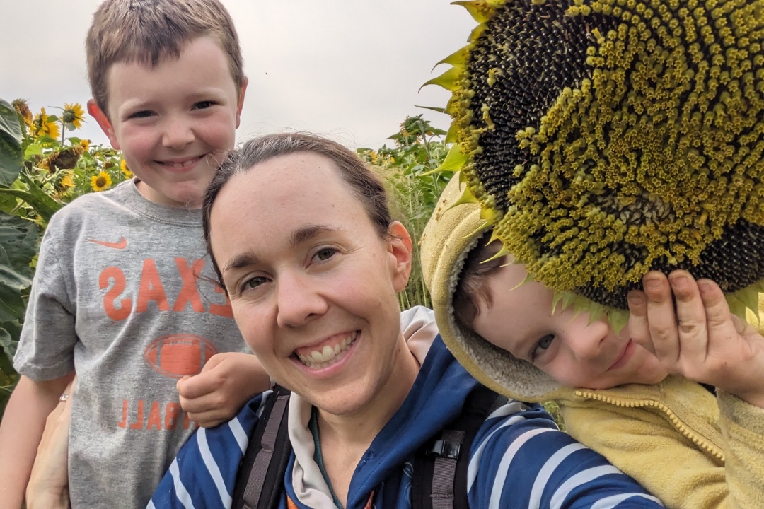 A brunette woman and two children smile amidst a field of sunflowers.