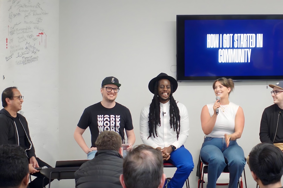 A panel of five people sit on stools behind a small audience in front of a blue screen with text reading "How I Got Started in Community."