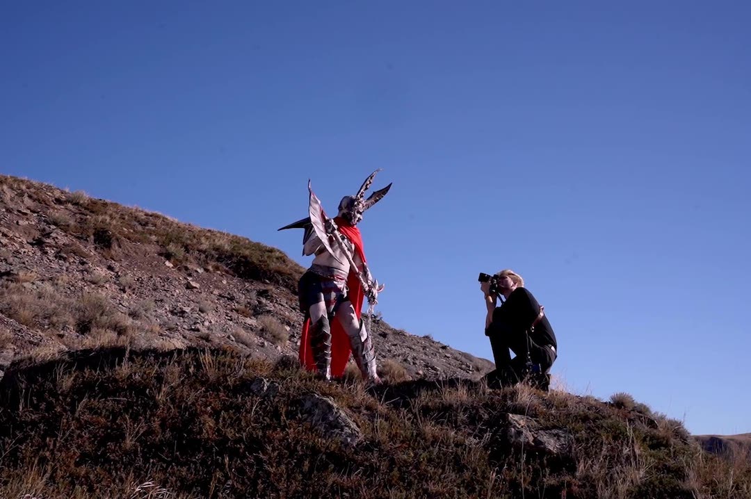 A drag performer poses near the summit of a butte, wielding a sword, while a photographer squats to the right and takes their photo.