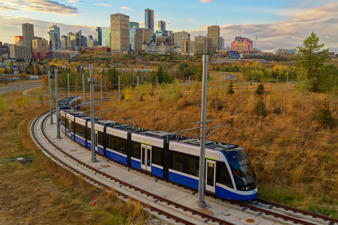 An LRT train runs along a track through a grassy field in the fall, with Edmonton's skyline in the background