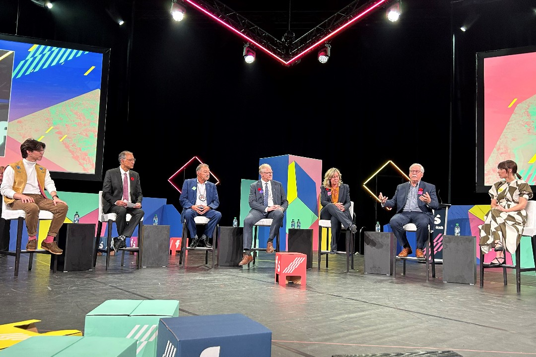 Six people sitting on elevated chairs on a stage listen to a seventh speaking