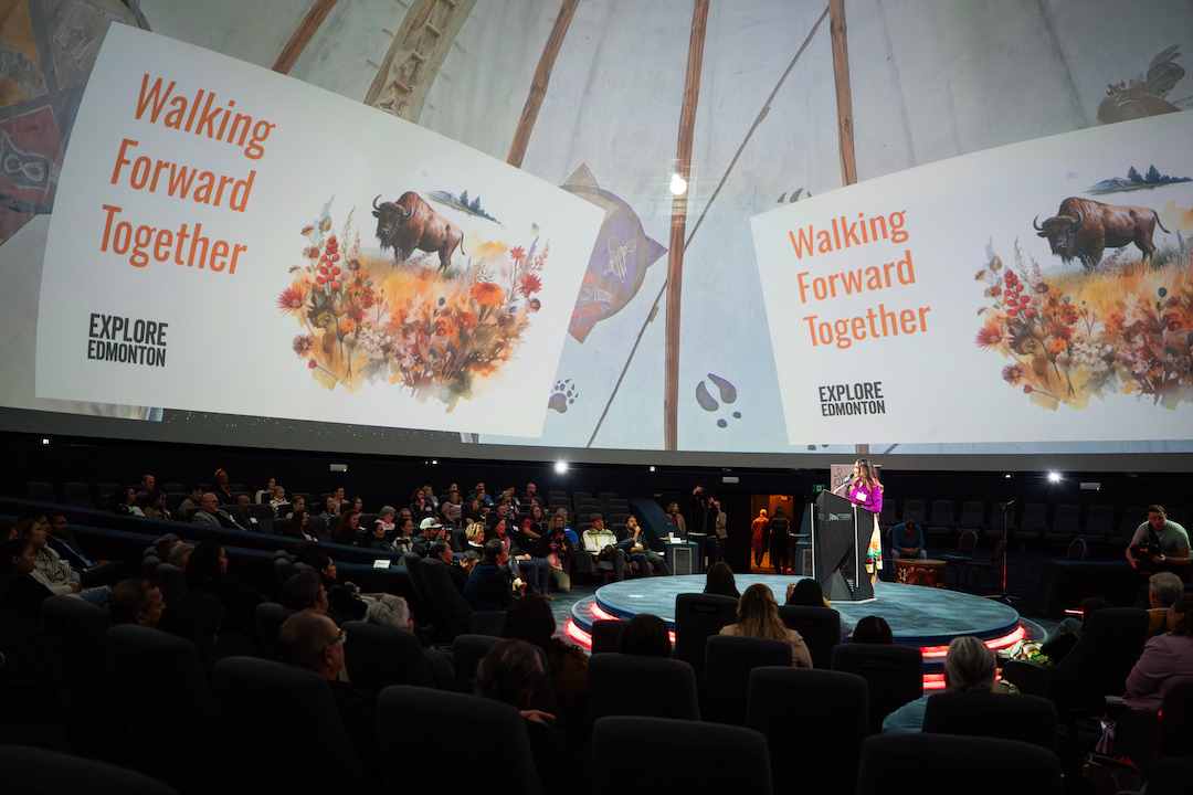 A woman stands at a podium on a circular stage, surrounded by an audience. Above them, two large panels read "Walking Forward Together" and "Explore Edmonton."