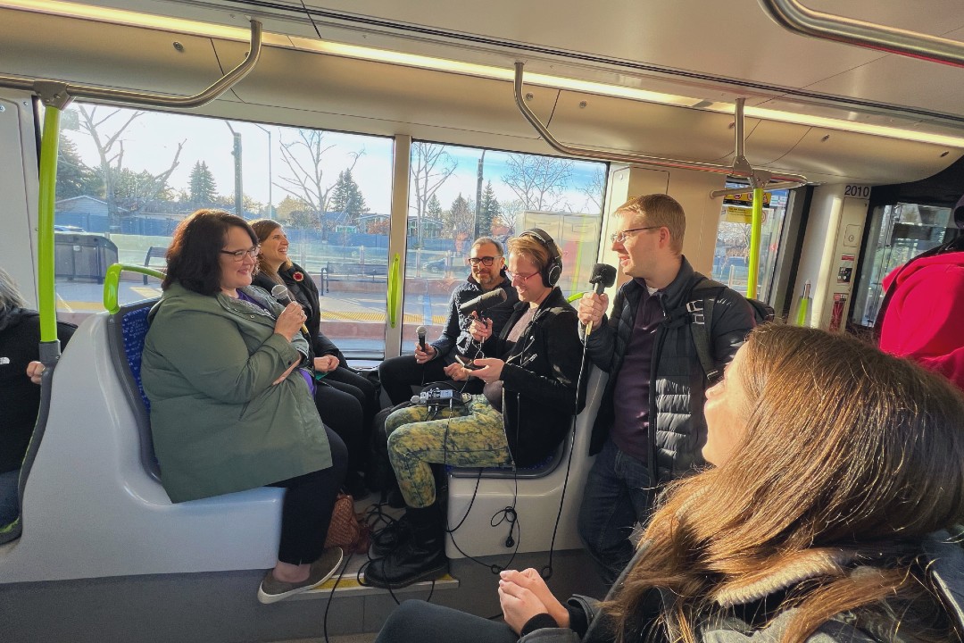 Several people hold podcasting microphones while riding the Valley Line Southeast LRT.