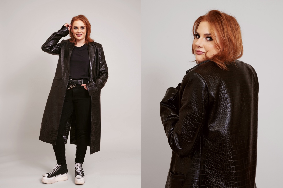 A woman with red hair poses in a long leather, crocodile-skin patterned trench coat and dark jeans.