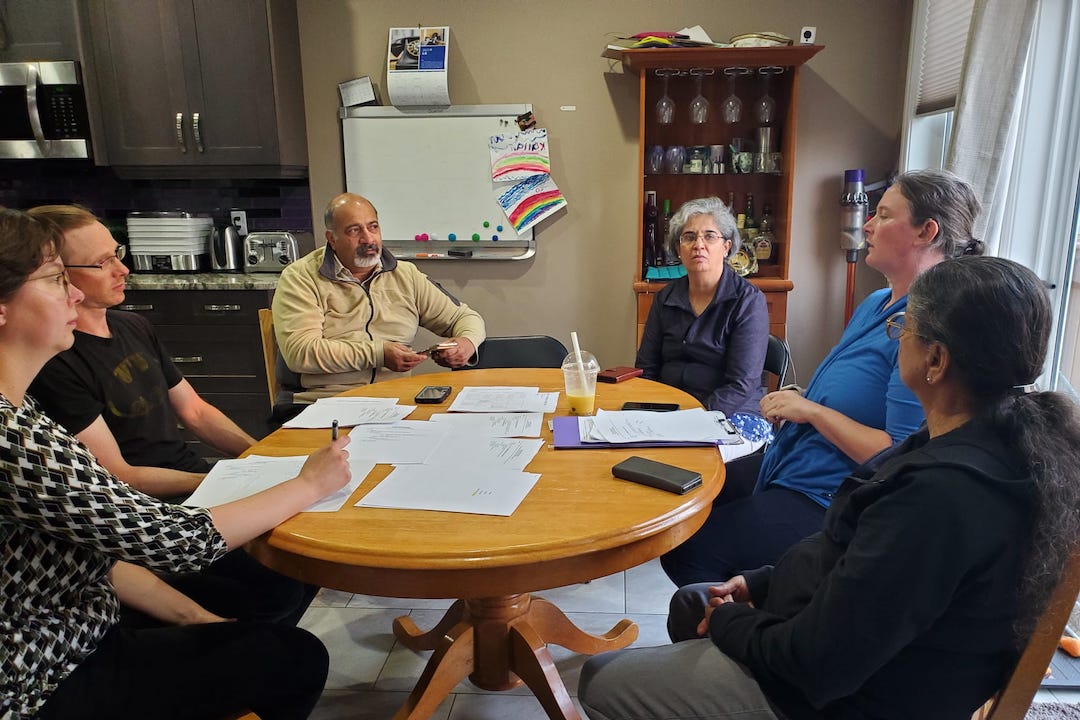 Six people gather around a table at the edge of a kitchen.