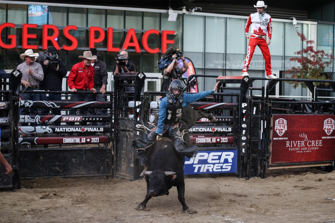 A bull rider atop a bucking bull with cowboys in the background cheering him on.