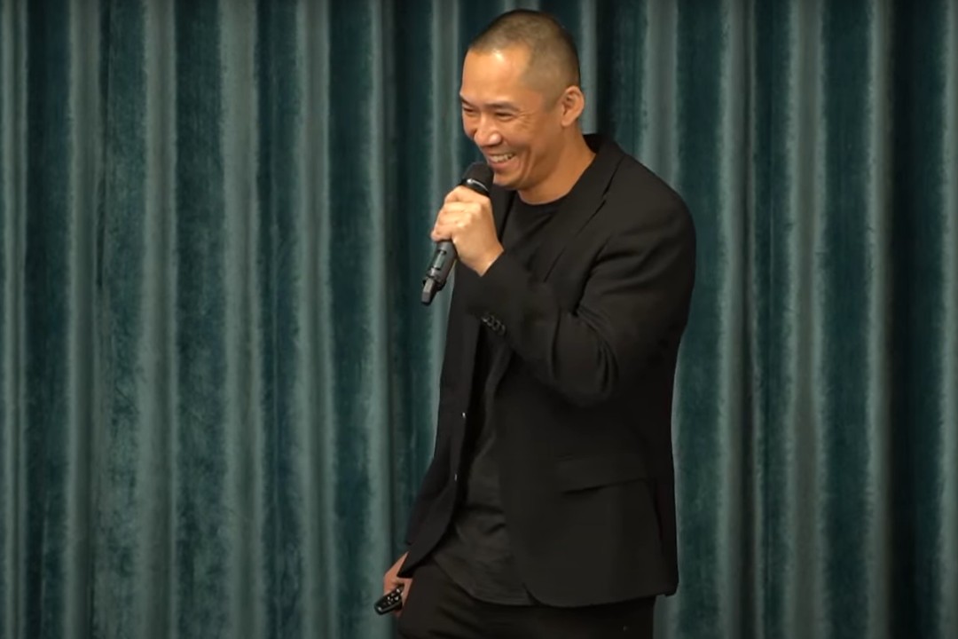 A man in a black suit smiles while speaking into a microphone.