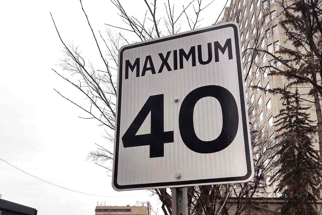 A street sign indicating a 40 km/h speed limit in front of leafless trees, a tall building, and an overcast sky.