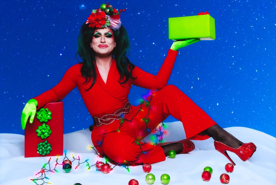A drag performer dressed in Christmas attire holds presents and poses amongst Christmas decorations.