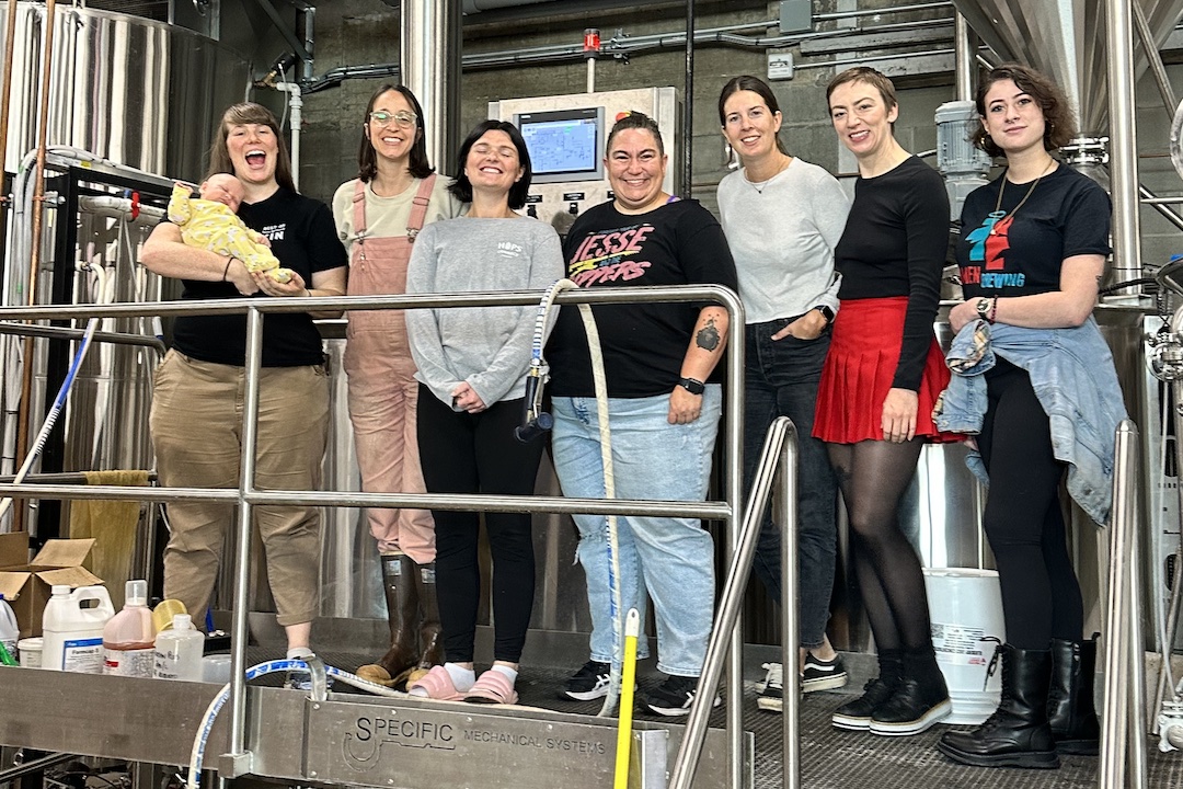 A group of seven women and non-binary people, plus a baby, pose inside a brewery.