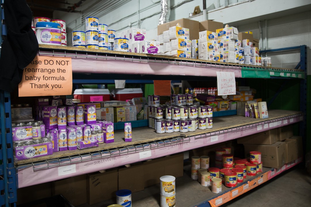An interior photo of Edmonton's Food Bank that shows shelves containing canned goods. A sign reads "Please DO NOT rearrange the baby formula. Thank you."