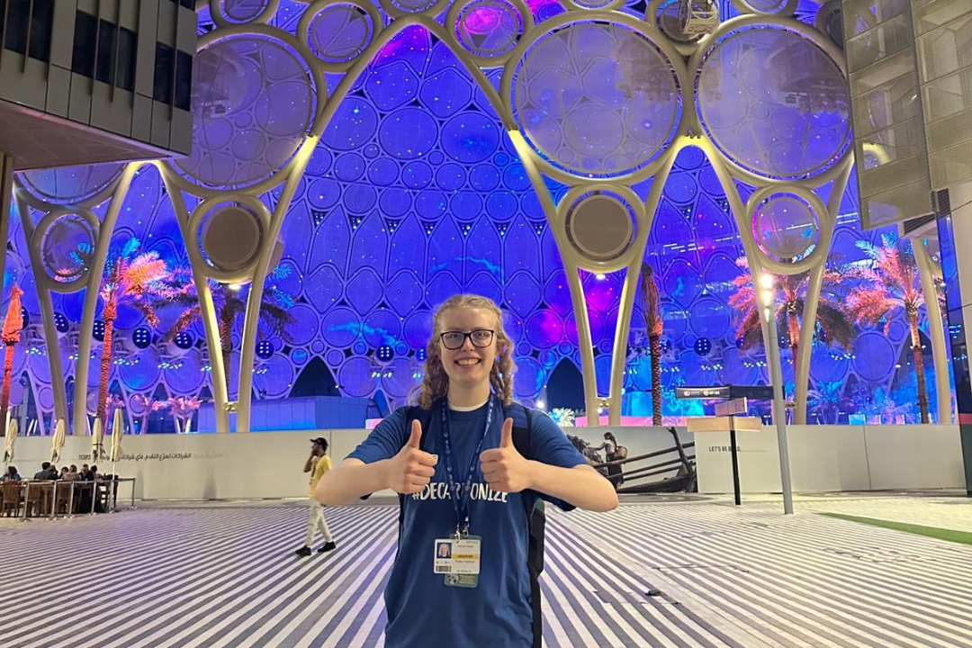 A person with blonde hair and black glasses gives two thumbs-up while standing before a domed pavilion lit with bright purples and blues.