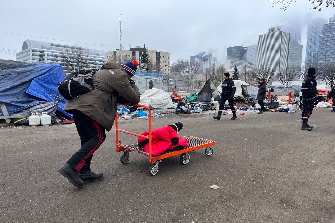 A person pushes a red cart with a blanket while in the background, police oversee an encampment eviction
