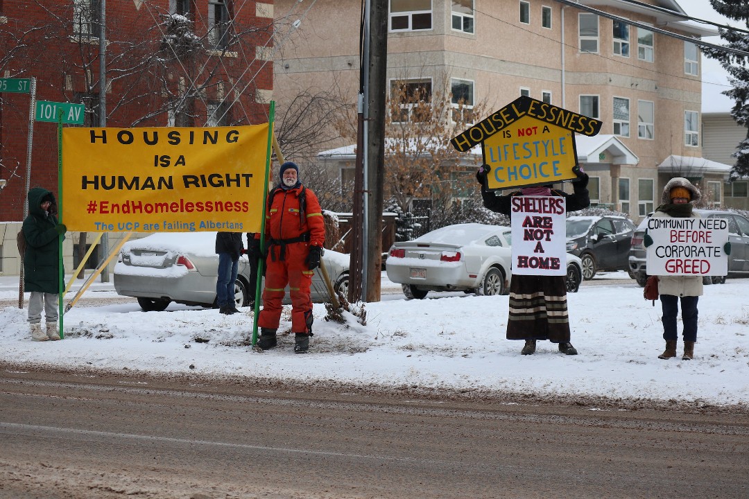 A group of people stand on the side of a road holding signs. One reads "Housing is a Human Right"; another reads "Shelters Are Not A Home."