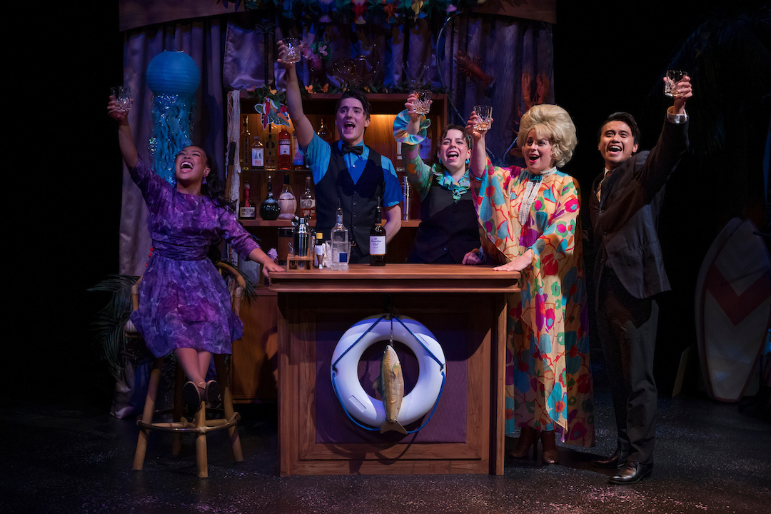 Actors pose in flamboyant costumes around a bar set on a theatre stage. Each is raising a glass.