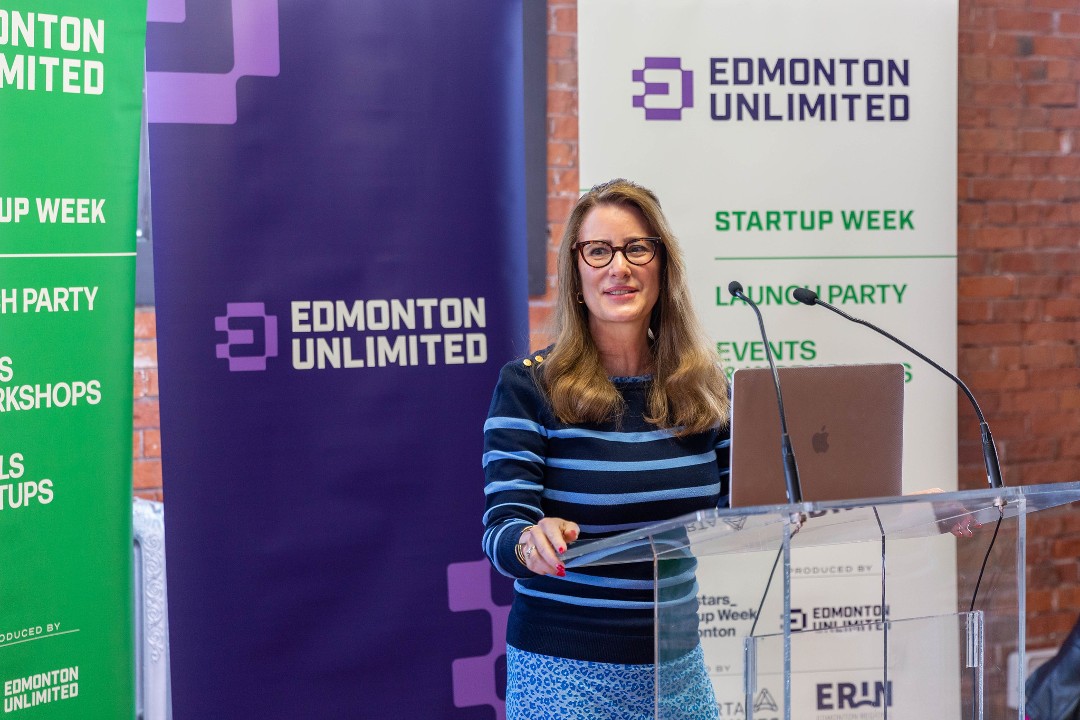 A person stands at a microphone while there is a sign that reads "Edmonton Unlimited" behind them.