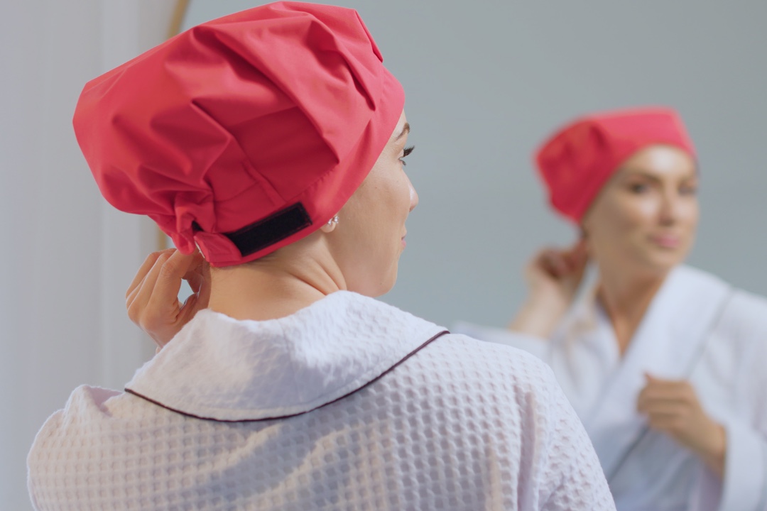 A woman in a white bathrobe wears a bright pink, adjustable shower cap and looks in the mirror