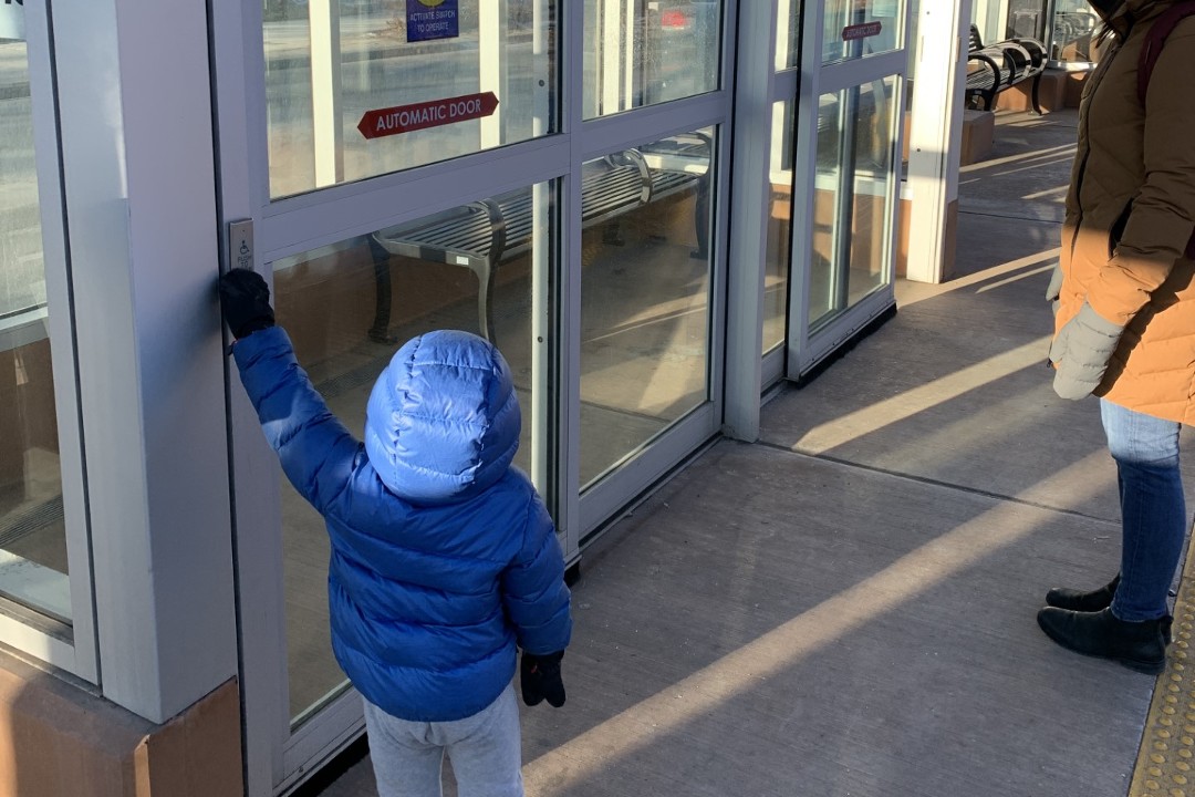 A toddler wearing a blue puffy coat presses a button to unsuccessfully make a shelter door open on the Valley Line LRT system.