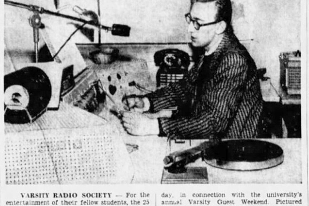A newspaper clipping of a photograph of a person sitting at a control board, surrounded by a turntable, and speaking into a microphone.