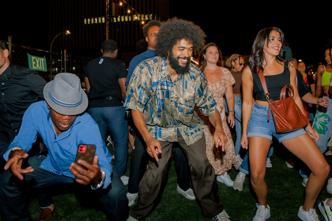 Smiling party-goers dance in a line beside a crouching man wielding a smartphone.