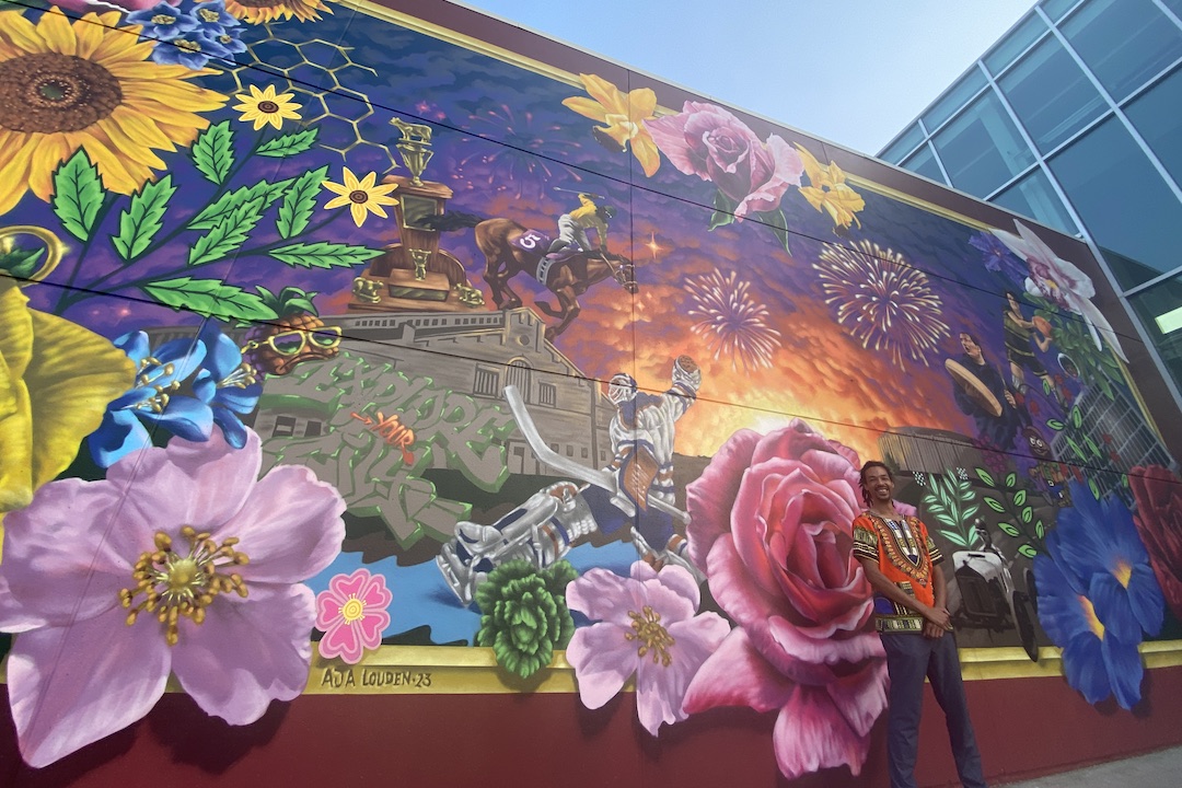 An artist poses in front of a large outdoor mural that depicts flowers, athletes, artists, and more.