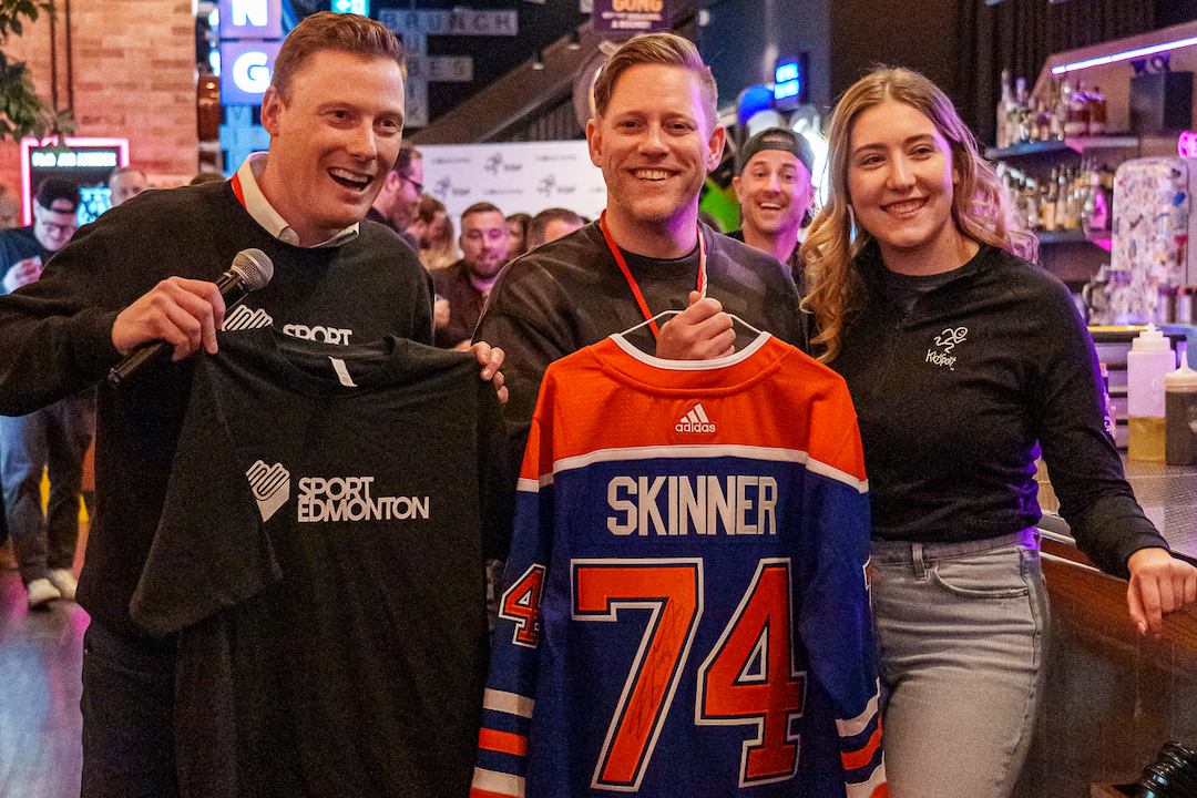 Three people gathered in a bar hold up a Sport Edmonton t-shirt and an Edmonton Oilers jersey for a player called Skinner.