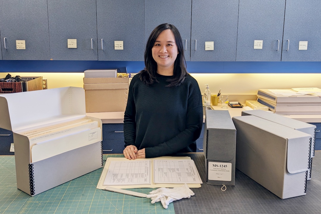A smiling person stands behind work tables, in front of cabinets, and surrounded by files.