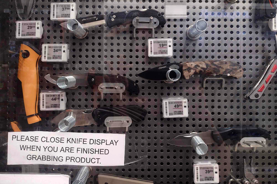 Knives for sale inside a retail display, with a note attached to the glass reading "PLEASE CLOSE KNIFE DISPLAY WHEN YOU ARE FINISHED GRABBING PRODUCT"