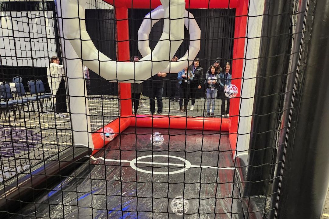 Young people stand behind an enclosed sports court. Inside it are drones encased in globe-shaped cages and two ring-shaped goalposts.