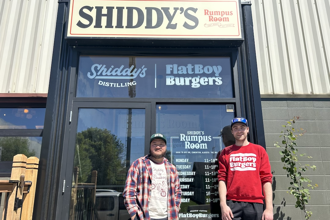 Two people wearing hats pose in front of a glass doorway with signage for Shiddy's Rumpus Room, Shiddy's Distilling, and Flat Boy Burgers.