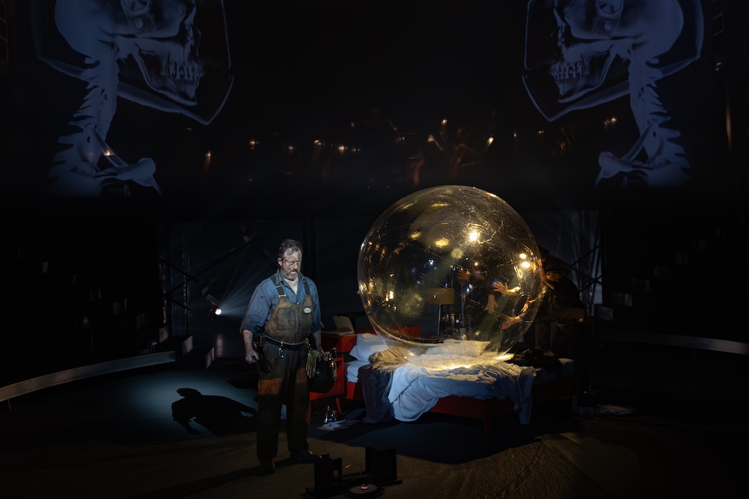 A person stands on stage next to a bed with a transparent orb on top