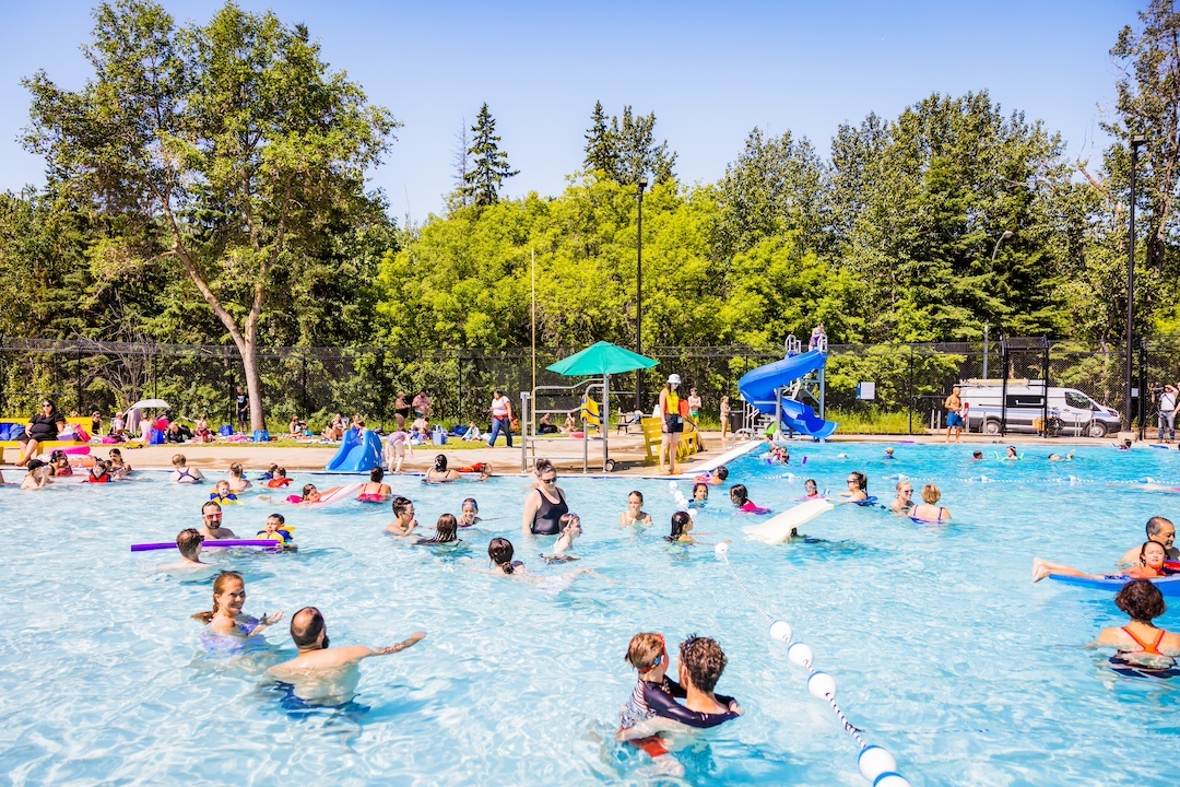 People swim in an outdoor pool. A slide and an umbrella can be seen in the background.