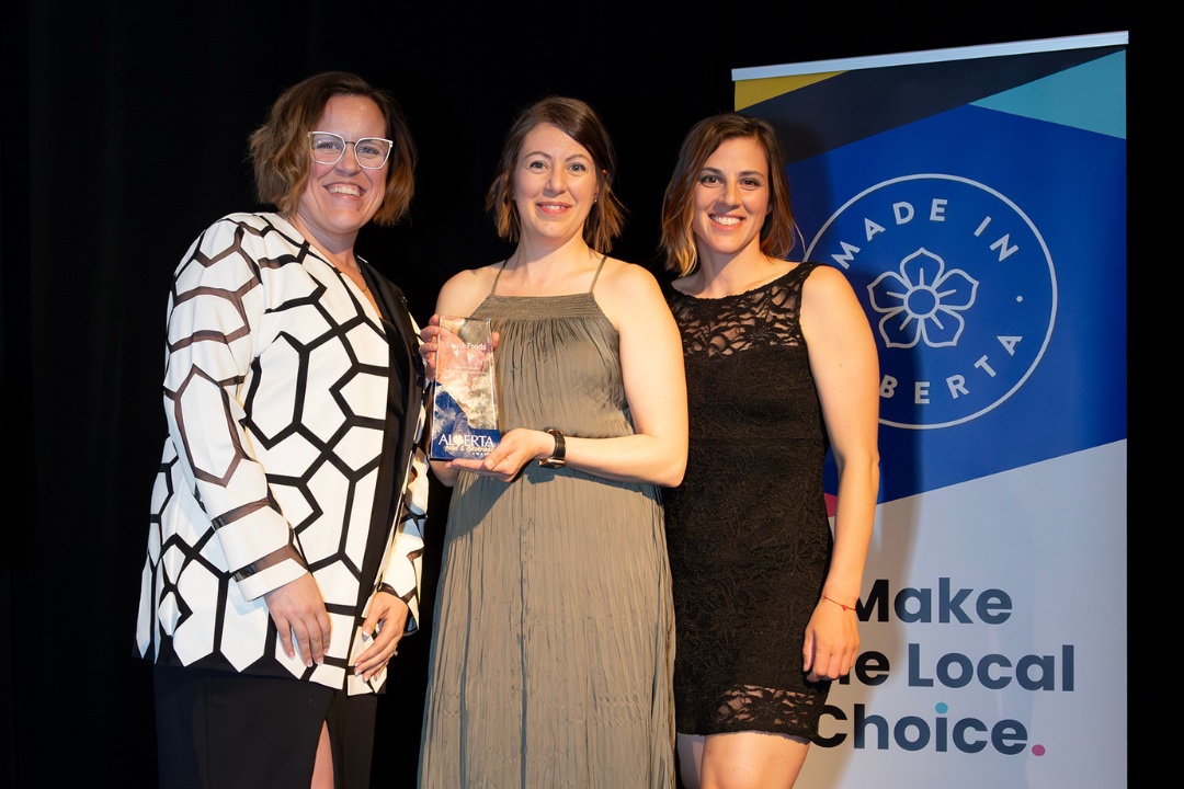 The person in the centre of a group of three holds an award. A banner behind them reads "Made in Alberta" and has other text that isn't entirely visible.