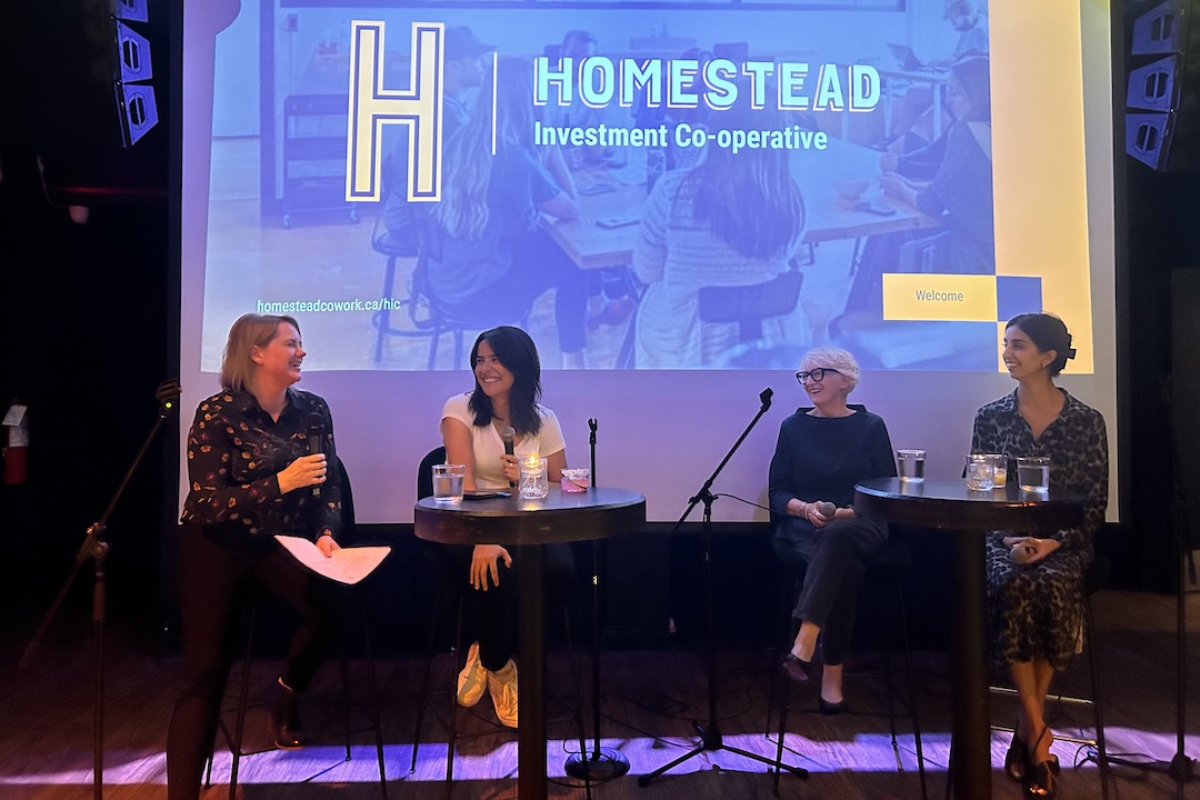 Four people sit at two cocktail tables with four microphones in front of a projector screen that reads "Homestead Investment Cooperative."