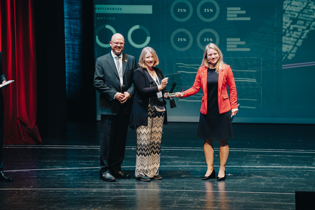 Three people on a stage. The one on the right hands an award to the one in the middle.