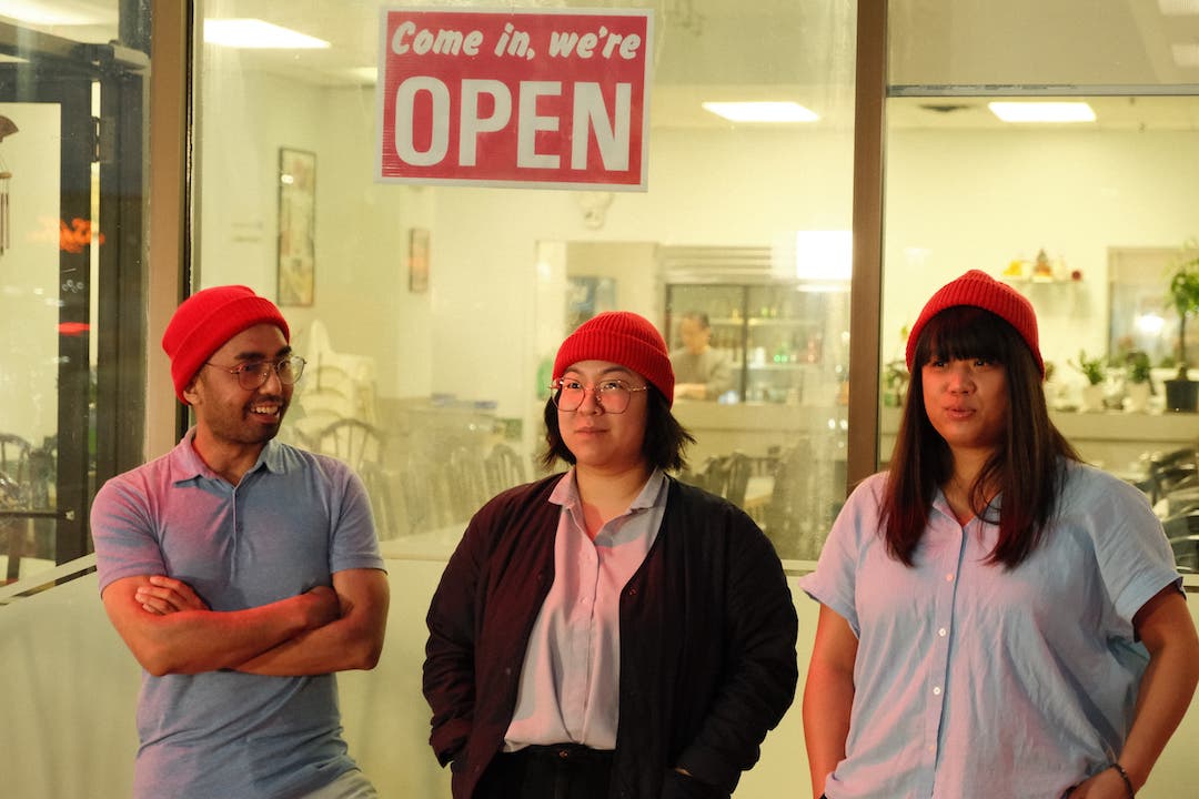 Three people wearing matching toques stand in front of a restaurant's front window. A sign behind them reads "Come in, we're OPEN."