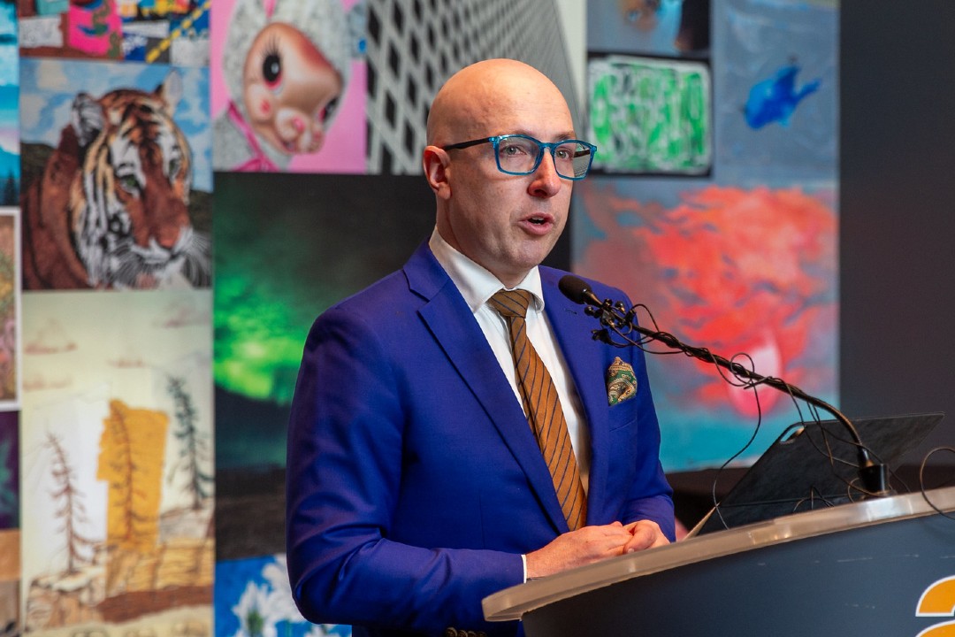 A person speaks at a microphone in front of an art-covered background