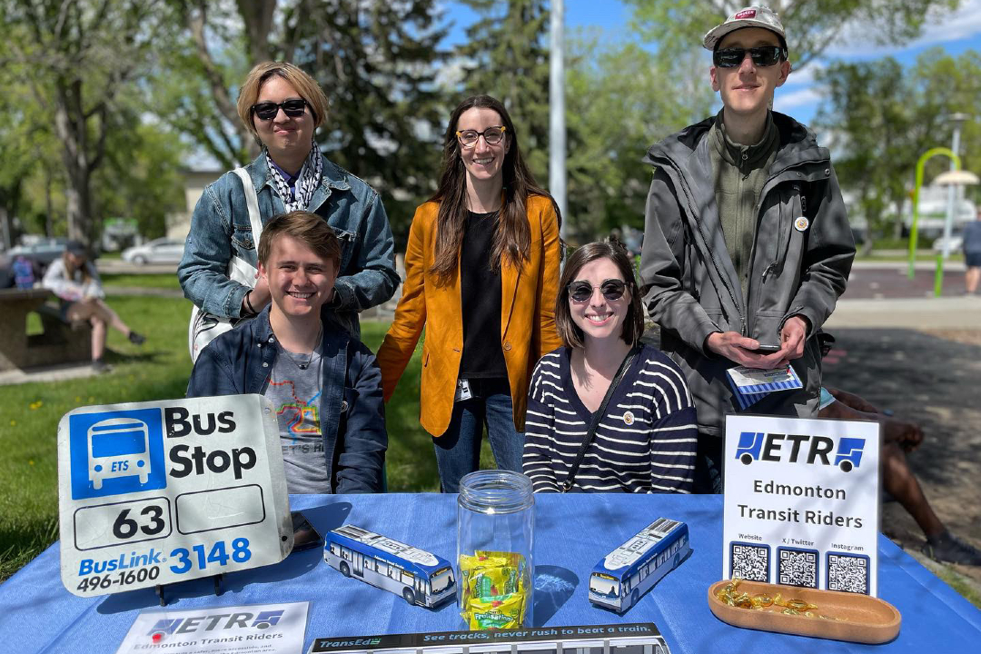 Five people gathered outdoors at a table covered in transit-themed paraphernalia, including a sign reading "Edmonton Transit Riders."