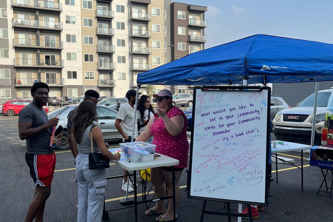 A woman standing behind a table under a tent talks to people in a parking lot. Next to her is a whiteboard asking for suggestions on how to activate the community.