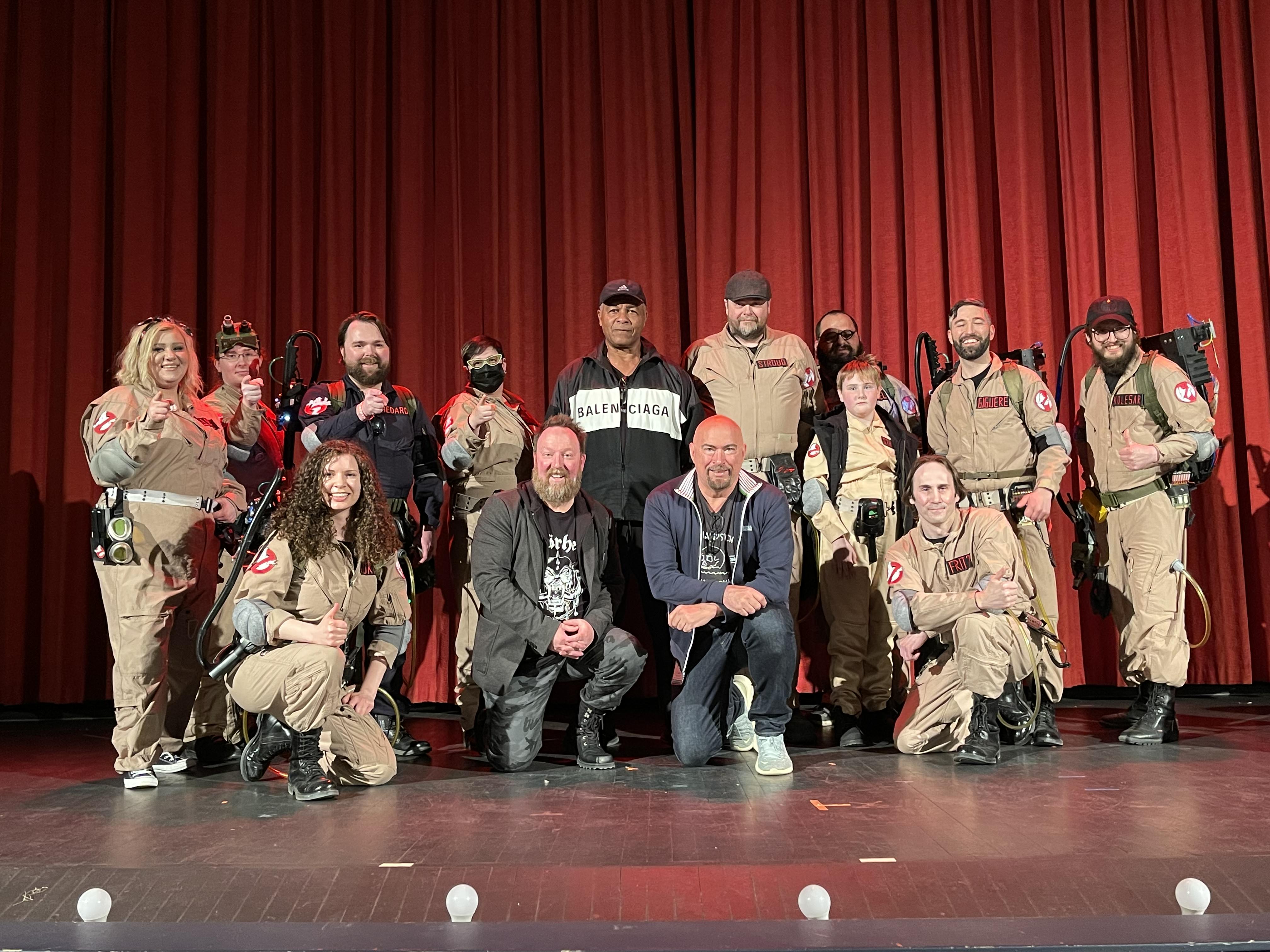 A group of people pose on a stage with red curtains behind them. Most are wearing Ghostbusters costumes, but two are kneeling in the centre of frame, and one man is stood among the costumed group with a Balenciaga hoodie on.