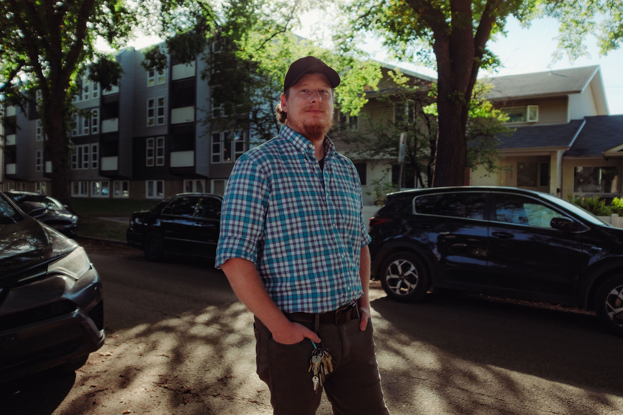 A man with a ring of trees dangling from his belt loop stands on a tree-lined, car-lined street with a low-rise apartment behind him