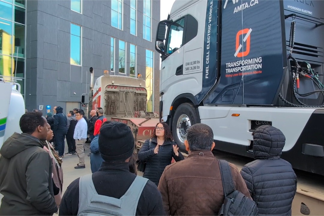 A group of people in winter wear gather outdoors between semi-trucks. One of the trucks is branded with the slogan "Transforming Transportation.