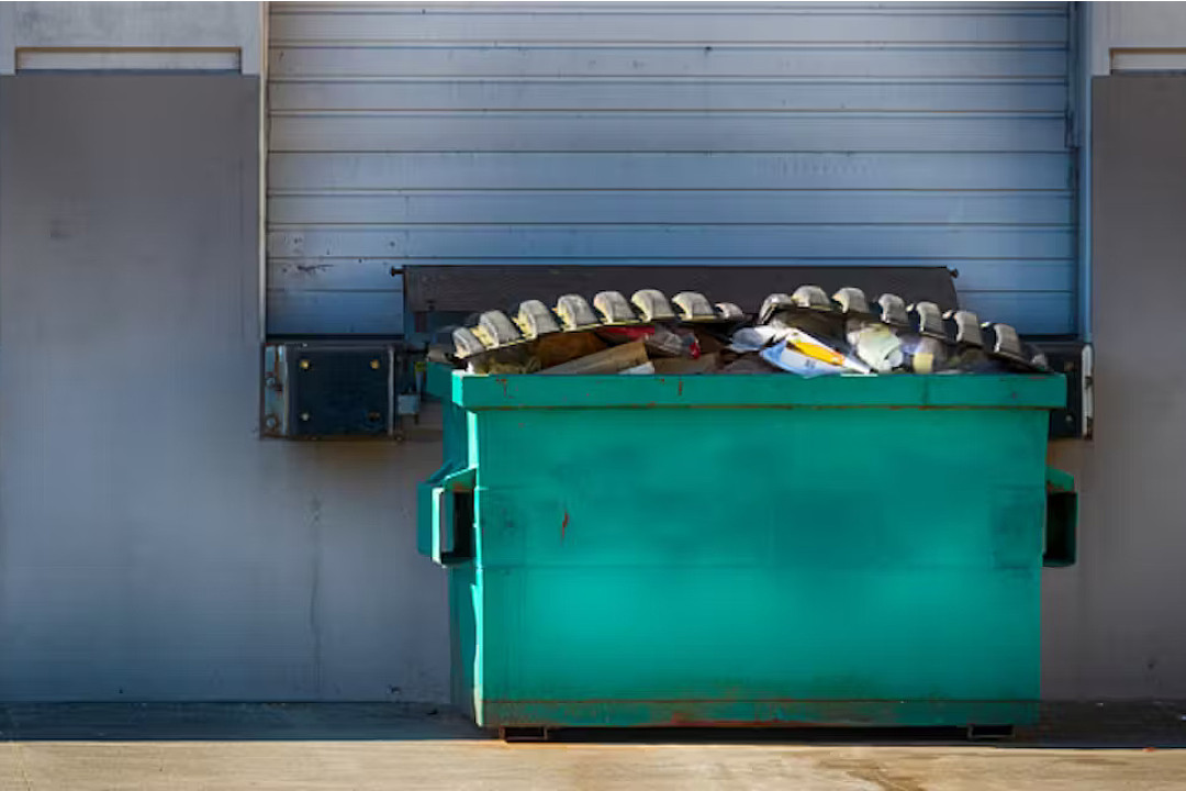 A dumpster, filled to the brim, next to the side of a building