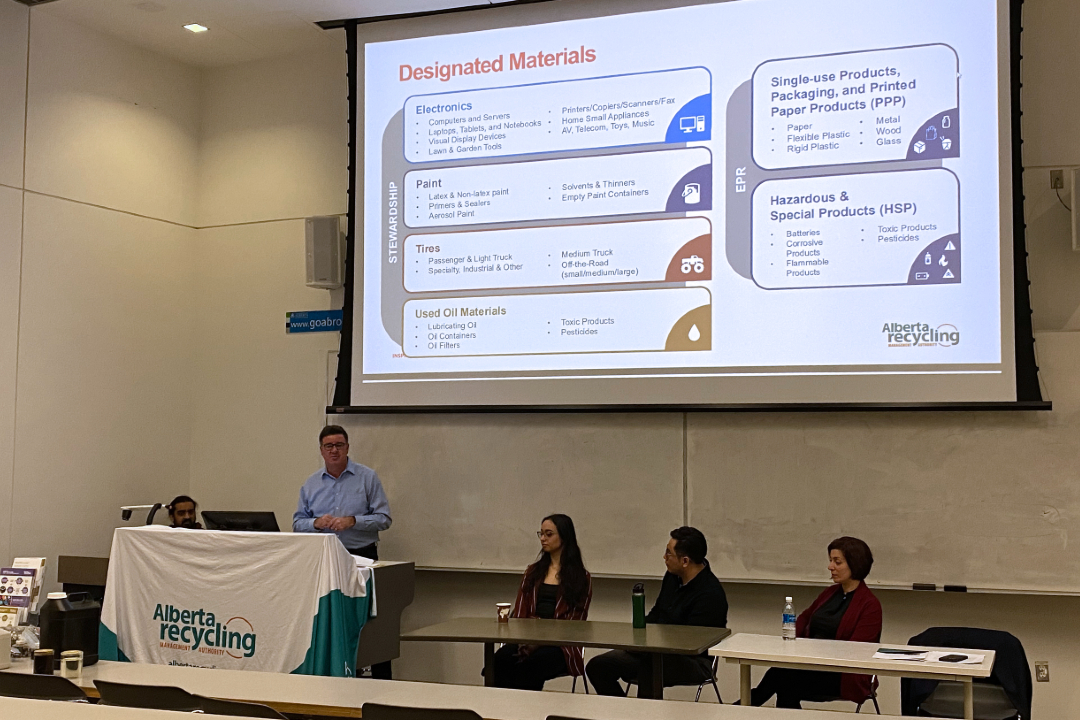 Staff, students, and guest speakers face students in a University of Alberta lecture hall. There's a banner on a podium with the logo for the Alberta Recycling Management Authority and a presentation projected on a screen.