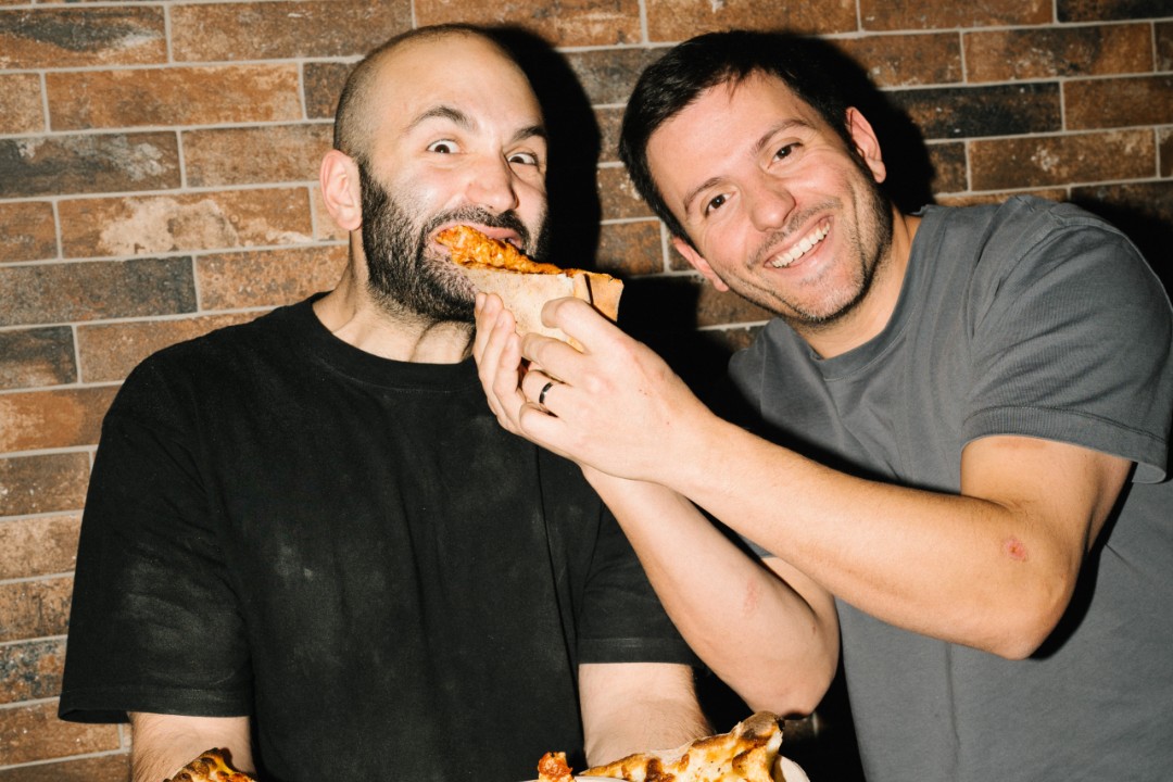 One man feeds another a slice of pizza.