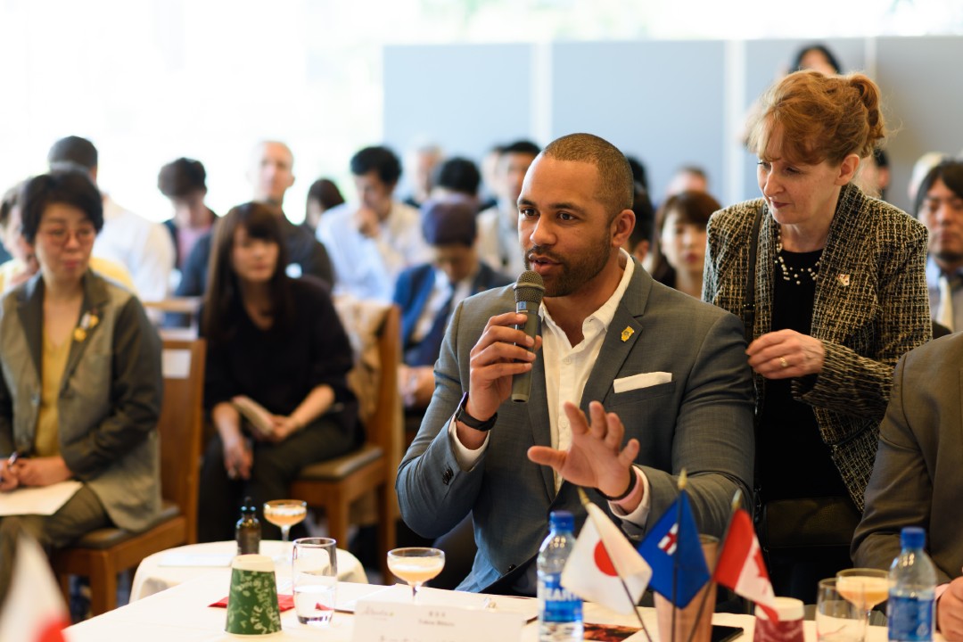 A man in a suit holds out one hand while using a microphone with the other while seated at a table.