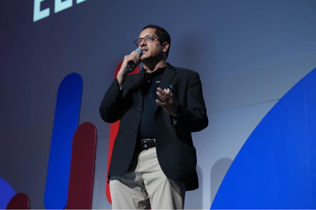 A man in business casual clothing speaks into a microphone on stage.