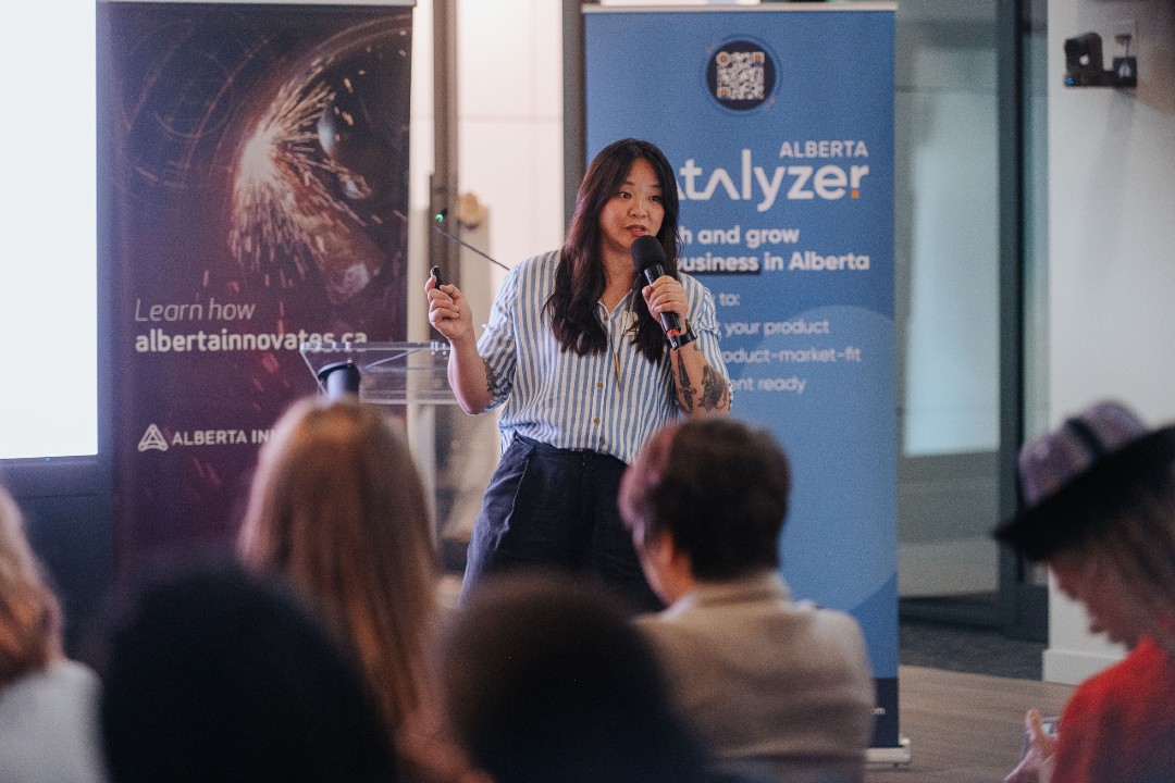 Linda Ha sets sights on helping freelancers avoid burnout and stay afloat A woman holds a microphone and remote and stands in front of banners for Alberta Innovates and Alberta Catalyzer.